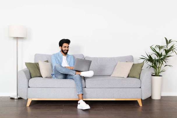 Θετικός χαρούμενος, όμορφος, χιλιετής Άραβας, που φοράει καθημερινά ρούχα και γυαλιά, χρησιμοποιώντας φορητό υπολογιστή, κάθεται στον καναπέ στο σπίτι. Ινδός ανεξάρτητος εργολάβος που εργάζεται σε απευθείας σύνδεση, αντίγραφο χώρου - Φωτογραφία, εικόνα