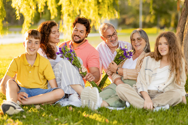 Happy caucasian multi-generation people rest, men give flowers to women, enjoy picnic in park. Relax, celebration holiday, meeting together outdoor, summer vacation, family relationship - Photo, Image