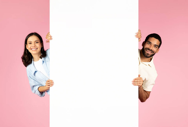 Pozitivní veselý krásný tisíciletý hispánský muž a žena pózující s vertikální prázdné bílé reklamní tabule, izolované na růžovém pozadí, prázdný prostor pro pěknou nabídku, řešení, text, mockup - Fotografie, Obrázek