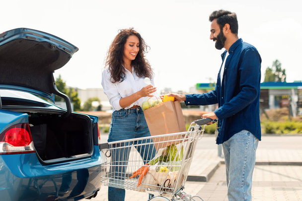 Married middle eastern couple putting paper bag full of healthy food into car, man helping wife, standing outdoors on parking near shopping mall - Photo, image