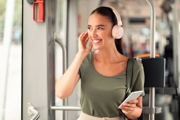 Hymyilevä matkustaja nainen, jolla on matkapuhelin ja kuulokkeet raitiovaunussa, nauttii upeasta musiikkisoittolistasta ja mukavasta ratsastuksesta nykyaikaisessa julkisessa liikenteessä, seisoo ja katselee sivuun - Valokuva, kuva
