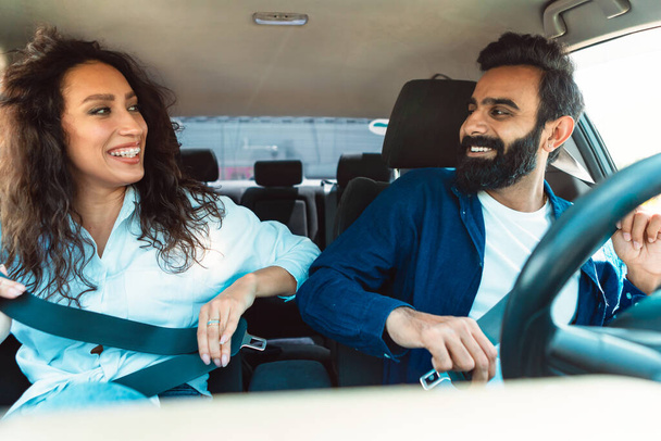 Opgewonden jonge echtgenoten uit het Midden-Oosten maken veiligheidsgordels vast, zitten in een nieuwe auto binnen, kijken elkaar aan en glimlachen. Auto passagiers veiligheid en auto verzekering service bieden concept - Foto, afbeelding