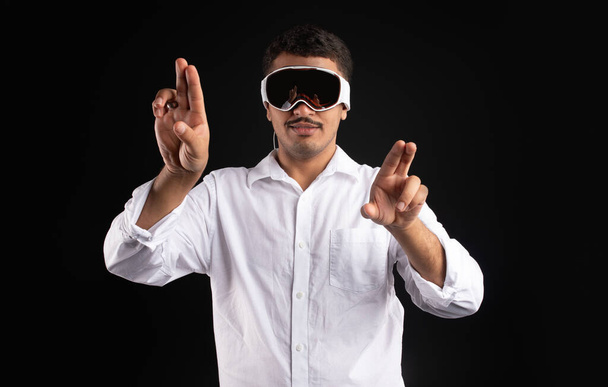 Latin man experiencing virtual reality in modern vision pro vr glasses, interacting with virtual screens gesturing hands and touching invisible touchscreen, black background - 写真・画像