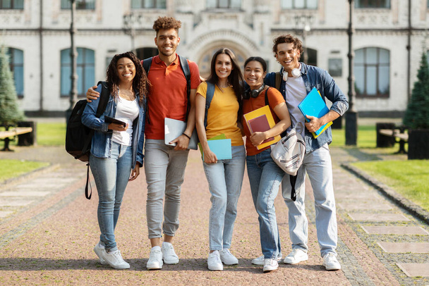 Portrait of happy international students posing outdoors near university building, cheerful young people smiling and embracing, multicultural friends with backpacks and workbooks standing outside - Photo, Image