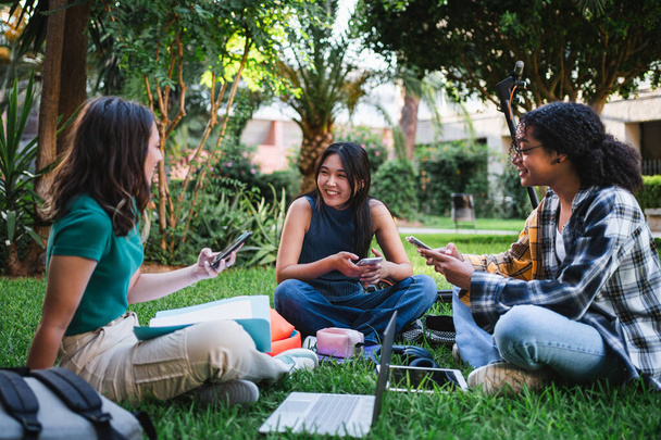 Drei lächelnde Universitätsstudenten, die auf dem Rasen sitzen, während sie sich vom Unterricht ausruhen. - Foto, Bild