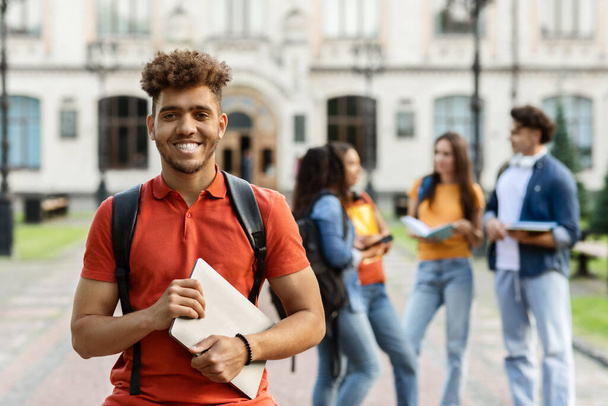 Έννοια υψηλής εκπαίδευσης. Πορτρέτο του χαρούμενου μαύρου φοιτητή που χαμογελά μπροστά στην κάμερα ενώ ποζάρει στην ύπαιθρο στην πανεπιστημιούπολη, τους συμμαθητές του και το πανεπιστημιακό κτίριο στο παρασκήνιο, επιλεκτική εστίαση - Φωτογραφία, εικόνα