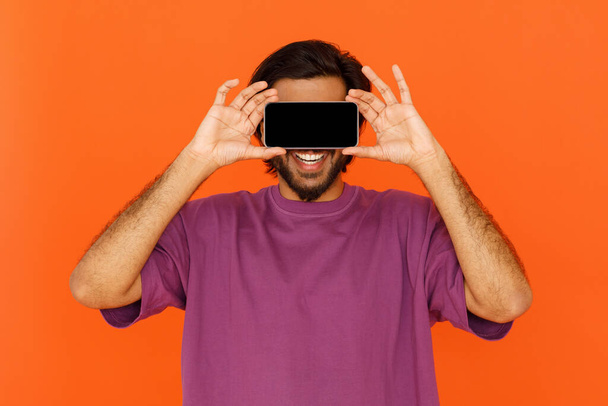 Communication, social media addiction, dating mobile app. Unrecognizable young man wearing casual purple t-shirt holding horizontal phone with empty black screen over eyes, orange background - Fotó, kép