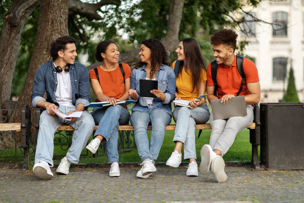 Verscheidene studenten vrienden zitten op de bank buiten op de campus, multi-etnische klasgenoten rusten samen, jonge mannen en vrouwen met behulp van digitale tablet en chatten, lachen en plezier hebben - Foto, afbeelding