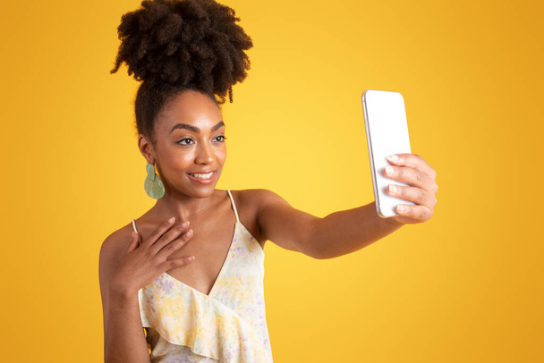 Счастливая молодая черная леди делает селфи на смартфоне, используя приложение социальных сетей, изолированное на желтом фоне, студия. Фотографии, объявления и предложения, образ жизни, видеозвонки, блоги и устройства - Фото, изображение