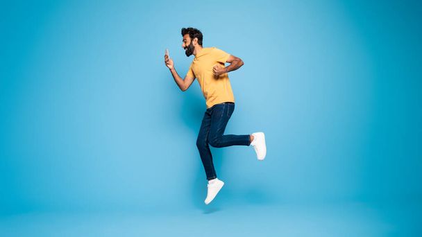 In voller Länge Aufnahme eines überglücklichen indischen Mannes, der springt, auf sein Smartphone blickt und auf blauem Hintergrund eine Geste macht, Panorama mit Kopierraum. Männchen gewinnt Online-Lotterie und erzielt damit Erfolg - Foto, Bild