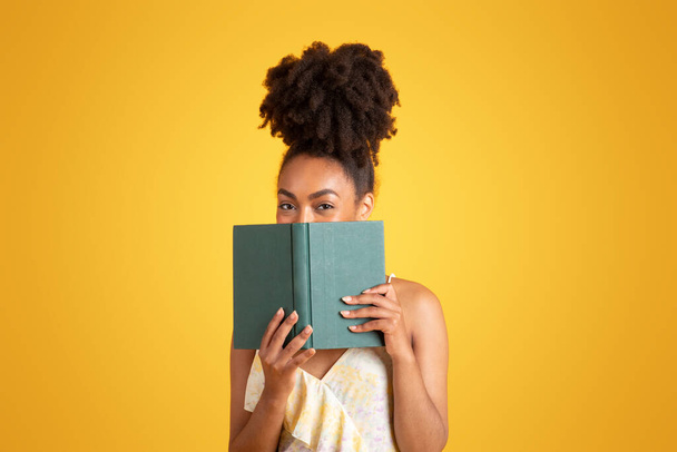 Cheerful young black woman reading book, has fun alone, isolated on yellow background, studio. Ad and offer, lifestyle, lady enjoy studies, education and hobbies in free time - Photo, Image