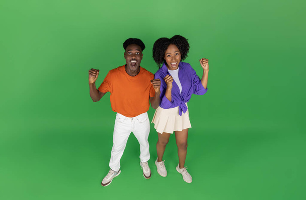 Glad african american millennial couple with open mouth celebrate win, success with hands, isolated on green studio background. Reaction to good news, emotions, sale, ad and offer, fun together - Photo, Image