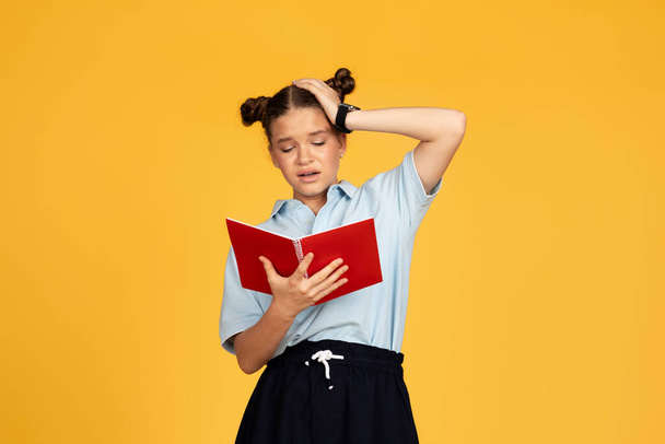 Tired unhappy teen european girl reads book, presses hand to head, isolated on yellow studio background. Problems with study, education at school, homework prepare for test emotions - Photo, image