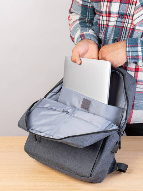 Un homme en chemise à carreaux met sa main sur un ordinateur portable gris dans un élégant sac à dos gris. - Photo, image