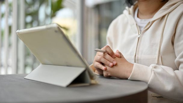 Imagen de cerca de una mujer freelancer asiática seria en un suéter acogedor trabajando en su trabajo en su tableta digital mientras está sentada en una cafetería. - Foto, Imagen