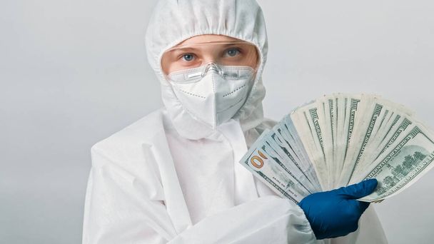 医療費だ。医者の収入だ。女性医療専門家女性でホワイトパイプブルー手袋ともにドルお金ファン孤立した上灰色の背景. - 写真・画像