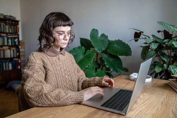 Chica adolescente enfocada usando gafas sentadas en la mesa escribiendo en el portátil, buscando información en la computadora mientras estudia en línea desde casa. Aprendizaje a distancia, tecnología y adolescentes - Foto, imagen