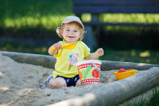 Happy toddler girl playing in sand on outdoor playground. Baby having fun on sunny warm summer sunny day. Active child with sand toys and in colorful fashion clothes - Фото, зображення