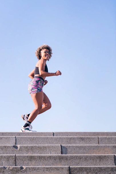 vertical photo of a happy african american woman running outdoors in a urban landscape, concept of sports and active lifestyle, copy space for text - Foto, Imagem