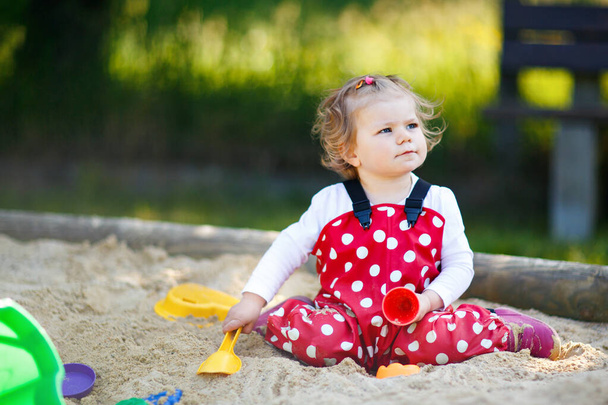 Roztomilé batole dívka hraje v písku na venkovním hřišti. Krásné dítě v červených gumových kalhotách baví na slunném teplém letním dni. Dítě s barevnými písečnými hračkami. Zdravé aktivní dítě venku hraje hry - Fotografie, Obrázek