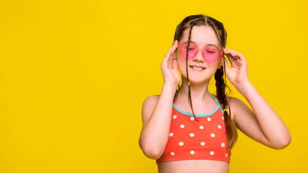 Boldog álmodozó lány élvezni nyáron, visel fürdőruha és a kezében rózsaszín napszemüveg elszigetelt sárga színű fal háttér stúdió - Fotó, kép