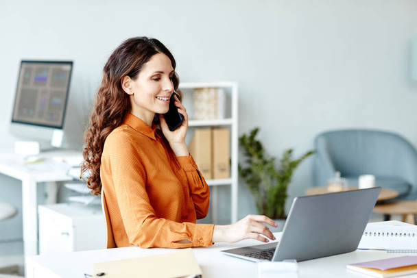 Νεαρή ενήλικη Καυκάσια επιχειρηματίας που κάθεται στο γραφείο της στο σύγχρονο γραφείο και εργάζεται με έγγραφα χρησιμοποιώντας φορητό υπολογιστή και μιλώντας στο τηλέφωνο - Φωτογραφία, εικόνα