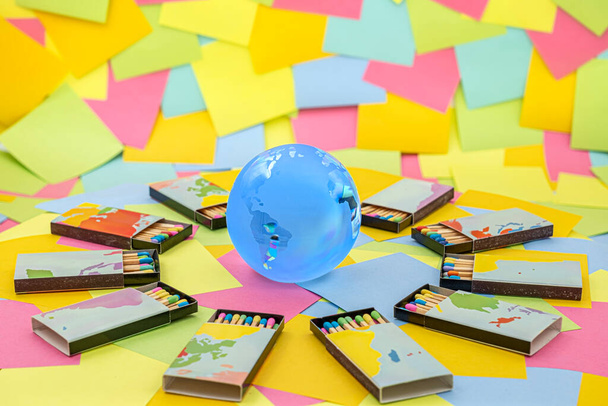 um globo de terra de cristal cercado por caixas de fósforos com paus coloridos em um fundo de papel multicolorido notas pegajosas - Foto, Imagem