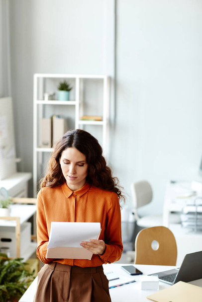 Tiro vertical de contable femenina con blusa naranja y falda marrón de pie en la oficina moderna comprobando los papeles financieros - Foto, Imagen