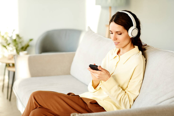 Jovem mulher caucasiana adulta usando fones de ouvido sentados no sofá em casa escolhendo música para ouvir no smartphone - Foto, Imagem
