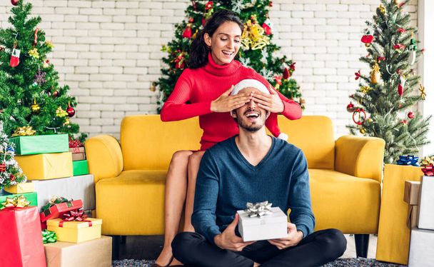 Romantic sweet couple in santa hats having fun decorating christmas tree and smiling while celebrating new year eve and enjoying spending time together.man giving gift box surprise to woman - Photo, Image