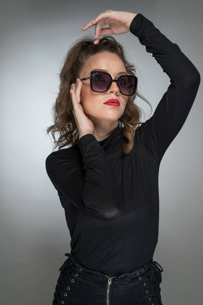 Woman with sunglasses  and black blouse against a grey background. Style beauty girl with red lips.Portrait of stunning young woman in sunglasses look into the camera - Photo, Image