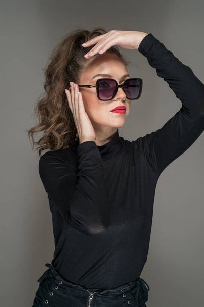 Woman with sunglasses  and black blouse against a grey background. Style beauty girl with red lips.Portrait of stunning young woman in sunglasses look into the camera - Photo, image