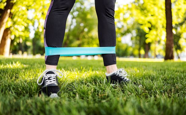 Girl with rubber elastic band doing leg workout outdoors on grass in park. Young woman exercising at morning with additional equipment for wellness - Valokuva, kuva