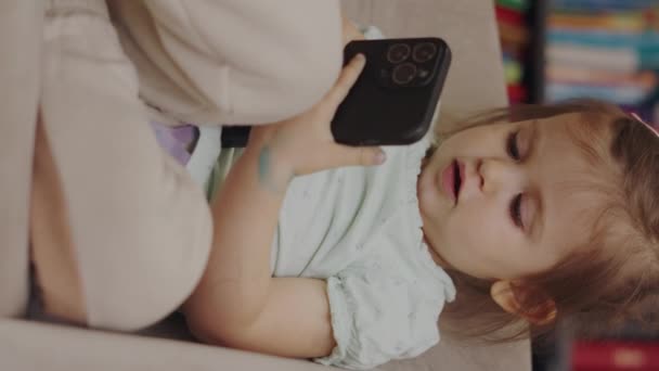 Bebek kanepede oturuyor, akıllı telefon ekranına dokunmaya dalmış, kanepede oyun oynuyor. İki yaşında bir çocuk. - Video, Çekim