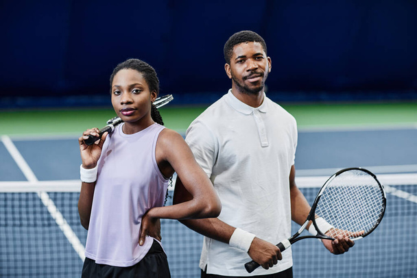 Afrikalı Amerikalı tenis oyuncularının belden yukarı portresi. Erkek ve kadın kameraya bakıyor. İç sahada raketlerle güvenle poz veriyorlar. - Fotoğraf, Görsel