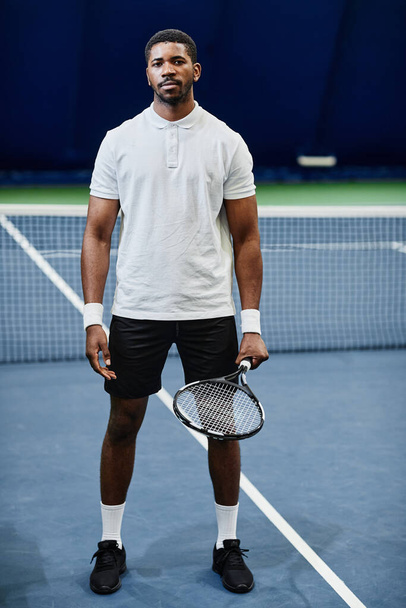 Full length portrait of African American tennis player looking at camera while posing confidently with racket at indoor court - Φωτογραφία, εικόνα