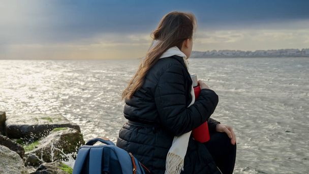 Νεαρή γυναίκα με μακριά μαλλιά κάθεται στους μεγάλους βράχους στη θαλάσσια ακτή και ατενίζει τον θυελλώδη καιρό πάνω από τον ωκεανό. Έννοια του τουρισμού, των ταξιδιών, του ταξιδιού, των διακοπών και των διακοπών το χειμώνα - Φωτογραφία, εικόνα