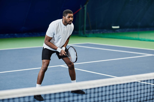 Πλήρες μήκος πορτρέτο του ευκίνητου μαύρου αθλητή που παίζει τένις στο εσωτερικό δικαστήριο, αντίγραφο χώρου - Φωτογραφία, εικόνα