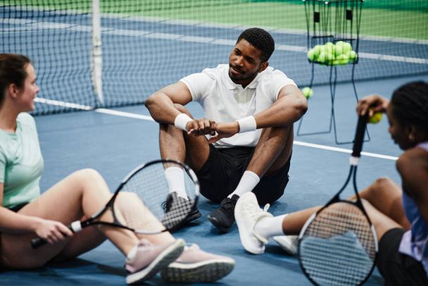 Grupo de personas que se relajan en la cancha de tenis cubierta y charlan mientras están sentados en el suelo - Foto, imagen