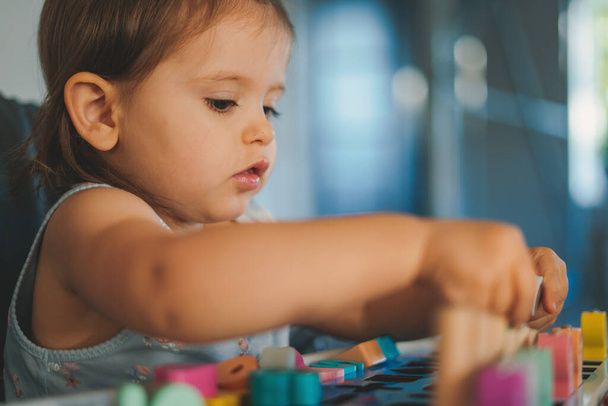 Πορτρέτο του παιδιού παίζει με ξύλινα σχήματα και αριθμούς και πολύχρωμα πλαστικά τούβλα στο τραπέζι. Το παιδί διασκεδάζει. Πρώιμη μάθηση. Ανάπτυξη παιχνιδιών - Φωτογραφία, εικόνα