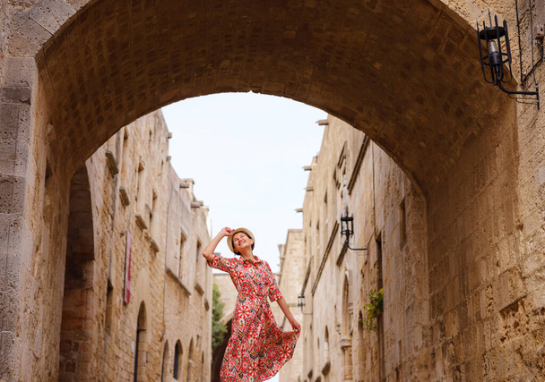Letnia wycieczka na wyspę Rodos, Grecja. Młoda Azjatka w etnicznej czerwonej sukience spaceruje ulicą Rycerzy Zamku Fortyfikacji. podróżniczka odwiedzająca południową Europę. UNESCO Światowe dziedzictwo UNESCO. - Zdjęcie, obraz