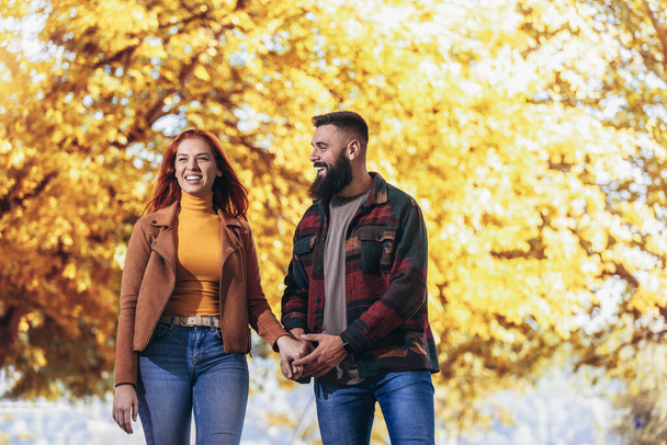 Genç mutlu çift el ele tutuşup sonbahar parkında yürürken birbirlerine bakıyorlar.. - Fotoğraf, Görsel