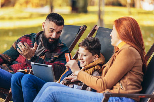 Joven familia disfrutando del tiempo en una cafetería al aire libre usando tableta digital para compras en línea. - Foto, Imagen