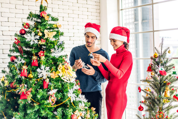 Ρομαντικό γλυκό ζευγάρι σε Σάντα καπέλα διασκεδάζοντας διακόσμηση χριστουγεννιάτικο δέντρο και χαμογελώντας ενώ γιορτάζει την παραμονή της Πρωτοχρονιάς και απολαμβάνοντας το χρόνο που περνούν μαζί τα Χριστούγεννα στο σπίτι - Φωτογραφία, εικόνα