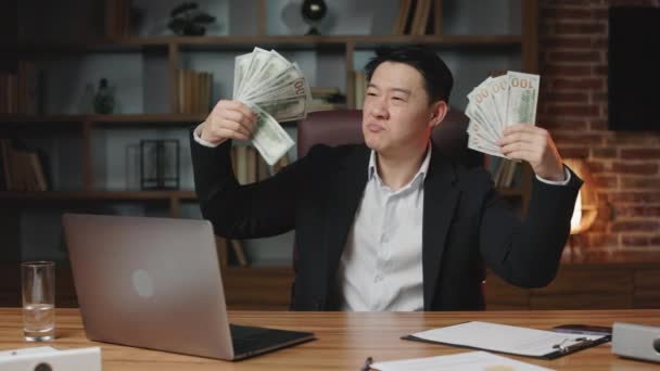 Proud business executive holding hundreds of dollars in both hands and fanning money in front of face. Dark-haired asian man enjoying prosperity sitting at desktop with remote computer at work. - Footage, Video