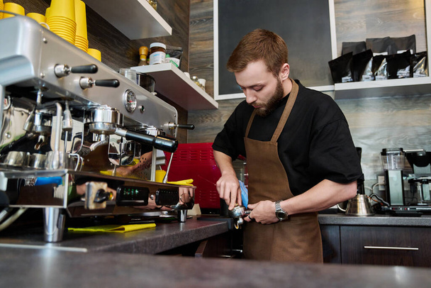 Male barista preparing coffee in coffee shop, bearded young man worker in an apron standing near coffee machine. Small business, food service occupation, coffee shop, staff, work concept - Φωτογραφία, εικόνα
