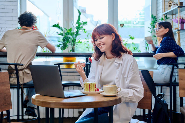 Female college student with laptop in cafe at table with cup of coffee and piece of cake. Internet online technology for leisure communication blogging learning chat, youth lifestyle concept - Photo, Image