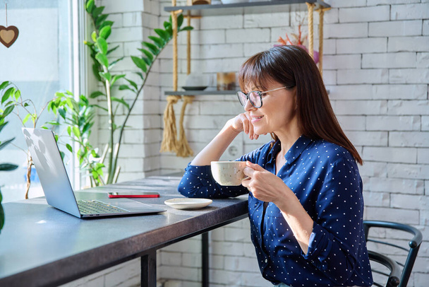 Middle-aged woman drinking coffee looking at laptop screen, sitting at table in cafe, coffee shop. Remote work, freelancing, blogging, chatting, online internet technology, mature people concept - 写真・画像