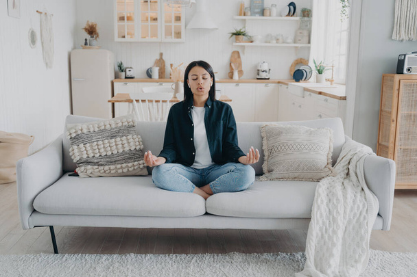 Serene vrouw beoefent yoga met mudra gebaar, ademend zittend op de bank in de woonkamer. Jonge vrouwelijke mediteren verlichten stress, ontspannen thuis. Gezonde levensstijl, emotie management. - Foto, afbeelding