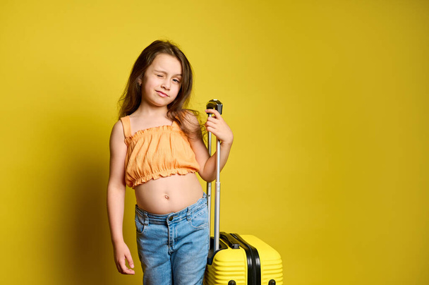 Cute Caucasian girl traveling abroad, going for weekend getaway, posing with yellow suitcase over yellow background, winking at camera. Children. Travel. Journey. Tourism. Summer. Holidays. Vacation - Photo, Image